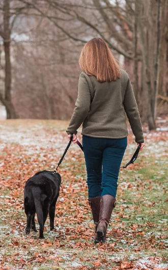 A veterinarian with shoulder-length hair, wearing a green jacket and brown boots, walks a black dog on a leash along a leaf-strewn path in a park on a chilly day.