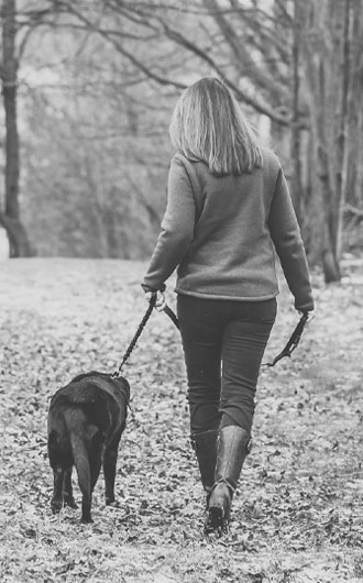 A woman with shoulder-length hair, possibly a veterinarian, walks a black dog on a leash along a leaf-covered path in a wooded area. The scene is in black and white, with both seen from behind.